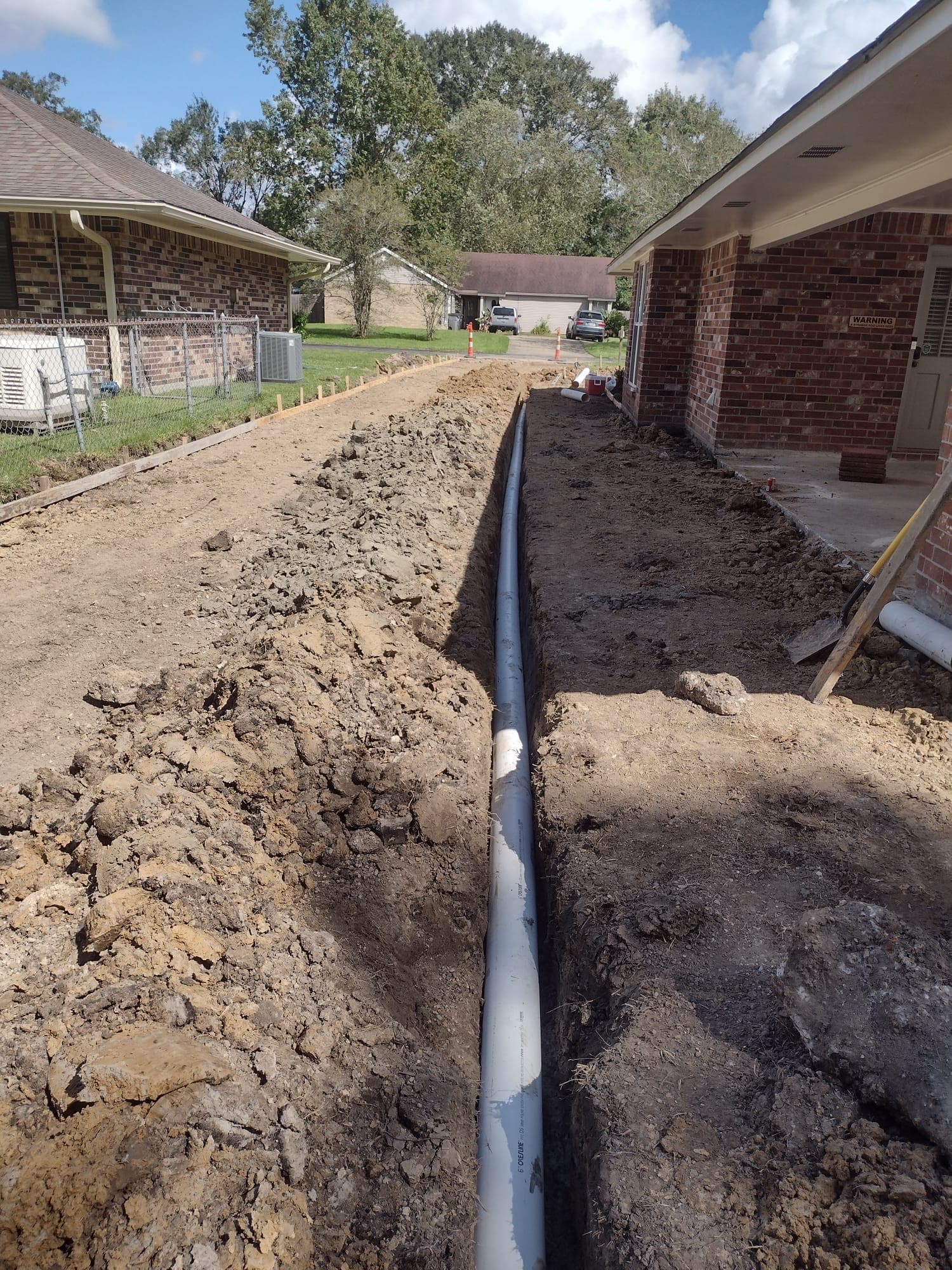 Drain pipe laid in a trench between two brick homes, with wood forms set along one side for the pour