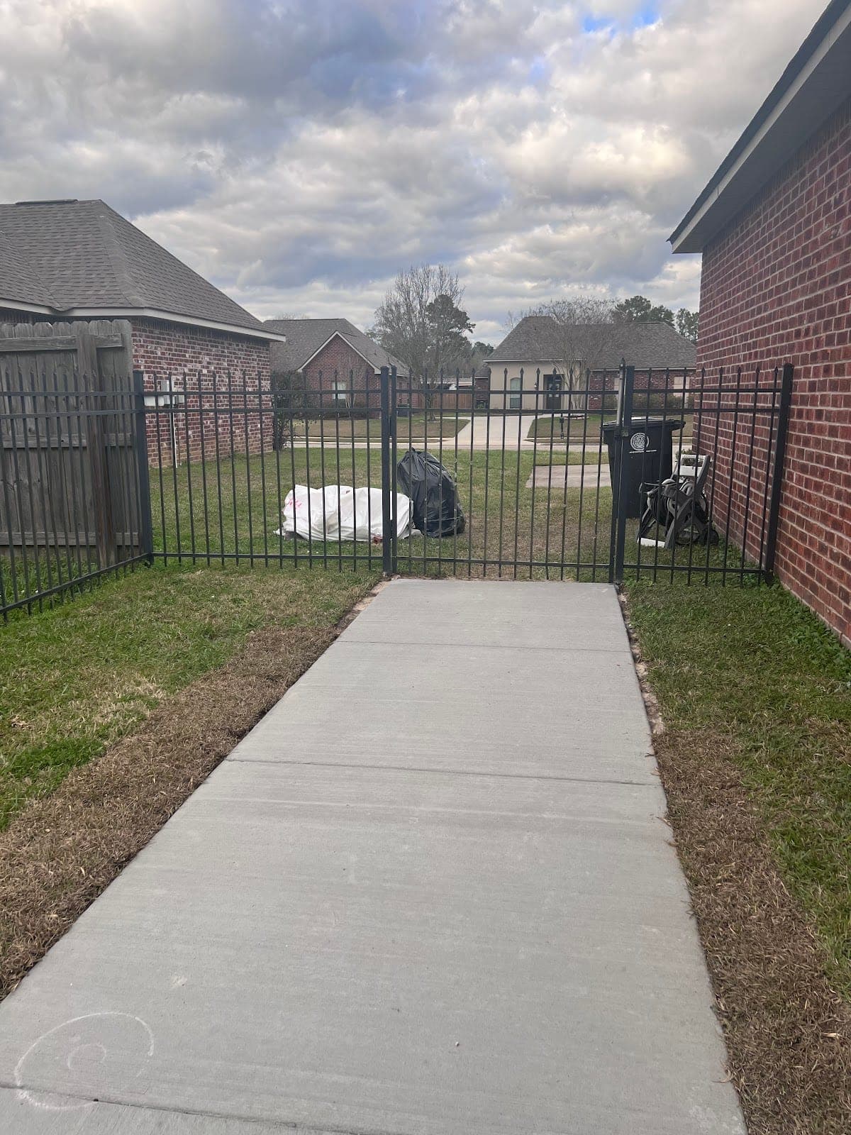 Finished concrete walk running between a brick home and a fence to a black iron gate