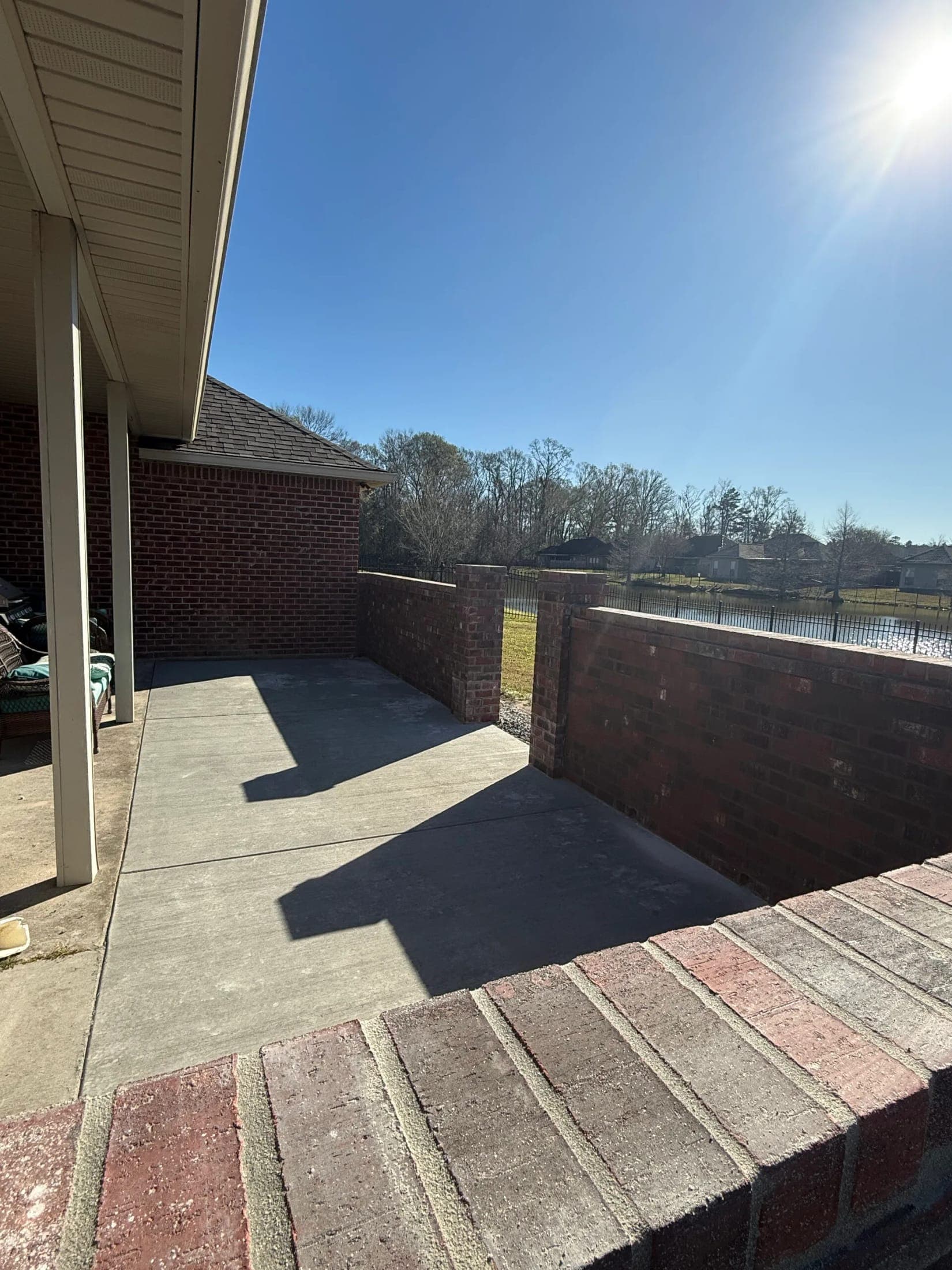 New concrete patio off a covered porch, enclosed by a brick half wall, with a pond beyond